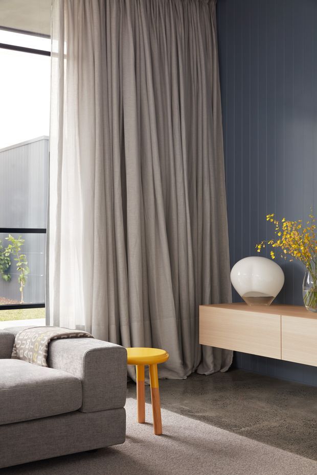 A refined contemporary living room corner with a large grey upholstered sofa, floor-to-ceiling taupe curtains, a dark blue-grey vertically panelled feature wall, and a floating light timber media console. A bright yellow side table, sculptural white table lamp, and vase of yellow foliage provide accents. Full-height glazing brings in abundant natural light, while a textured neutral carpet and dark polished-looking floor contribute to a warm, sophisticated atmosphere.