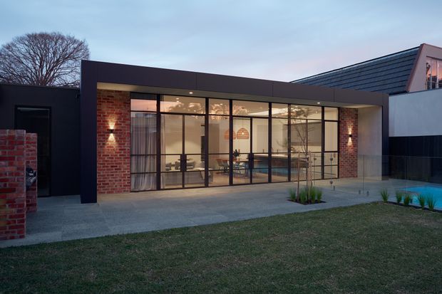 Contemporary single-storey residential extension viewed from the garden at dusk. The dark flat-roofed pavilion has extensive black-framed floor-to-ceiling glazing, exposed red-brick piers, and a broad concrete-paved terrace. A warmly lit dining and living area is visible through the glass, while a small pool edge appears at the right.