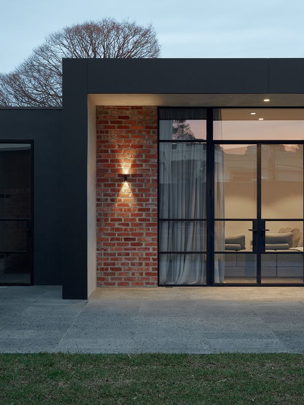 Contemporary residential facade at dusk, featuring dark charcoal cladding, exposed red brickwork, and a large black-framed glazed opening with double doors. Warm interior lighting reveals a living room beyond the glazing. A concrete forecourt and lawn sit in front, with a mature deciduous tree visible behind the flat-roofed structure.