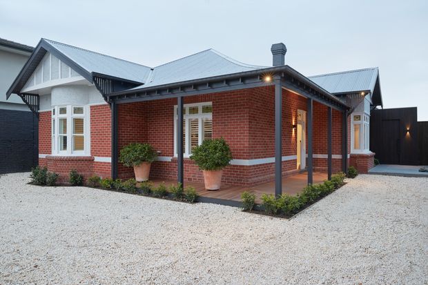 Single-storey red-brick residence with a heritage-inspired gabled roof form, projecting bay windows, white rendered detailing, and dark timber or metal verandah posts and trim. A covered verandah wraps the front corner, with low garden beds, potted shrubs, pale gravel paving, and a dark boundary fence. The exterior is photographed in soft overcast light.