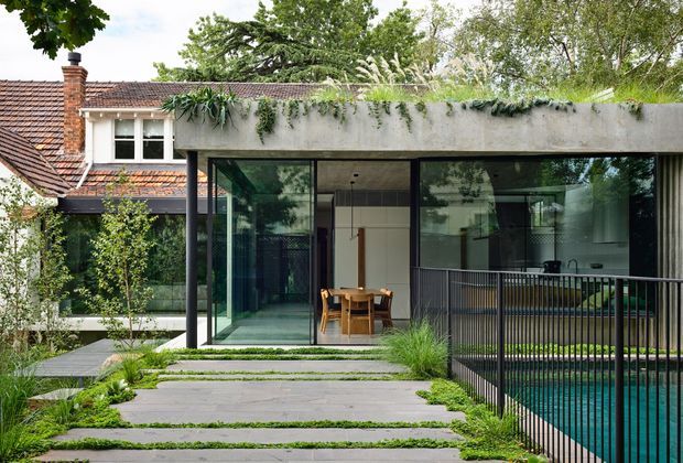 Exterior view of a contemporary flat-roof extension adjoining an older tiled-roof brick house. The extension has broad floor-to-ceiling glazing, dark slender framing, a planted roof edge, and a covered outdoor terrace overlooking a fenced swimming pool. Stepping-stone concrete paving with planted joints leads through a landscaped garden to the glazed dining area.