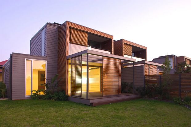 Exterior view of a contemporary multi-level residential development with box-like forms, warm horizontal timber-look cladding and extensive horizontal privacy screening. The homes feature recessed balconies, dark-framed glazing, projecting roof planes, and a glazed ground-floor outdoor room or terrace. A lawn and planted boundary sit in the foreground.