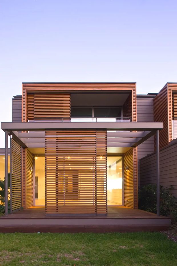 Front exterior view of a contemporary multi-level residence with extensive horizontal timber screening, dark horizontal wall cladding, a projecting flat-roofed canopy, recessed entry, upper balcony, and warm interior lighting visible through the screened facade. The home is set behind a small lawn and landscaped edges.