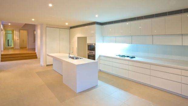 Bright, spacious contemporary kitchen with a large white island, integrated white cabinetry, concealed appliances, a gas cooktop, and a long illuminated benchtop. Pale tiled flooring and recessed ceiling lighting create a clean, minimalist appearance, with a stepped entry or hallway visible beyond the kitchen.