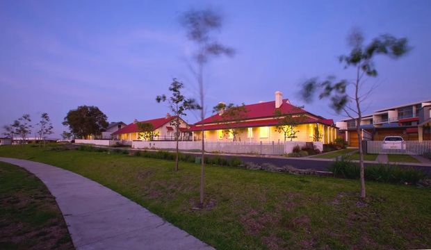 Dusk exterior view of a low-rise residential building with a long, single-storey form, bright yellow illuminated walls, red hipped roof, covered veranda, and white fencing. A concrete pedestrian path and landscaped lawn occupy the foreground, with neighbouring residences visible to the right.