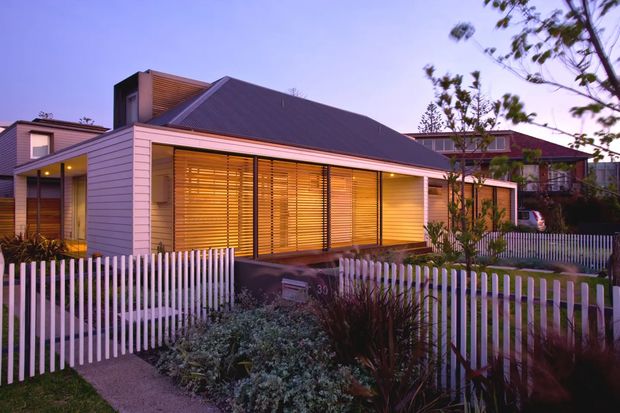 Twilight exterior view of a contemporary single-storey home with a dark pitched roof, pale horizontal weatherboard cladding, extensive timber-look louvred window screens, and a white picket-style front fence. A landscaped front garden and entry path sit in the foreground, with neighbouring homes visible behind.