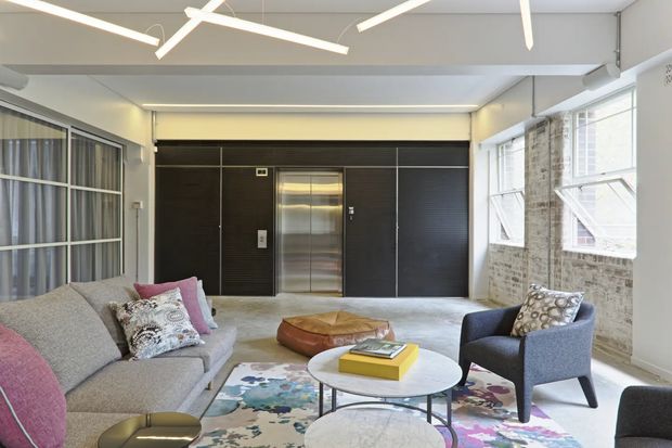 Bright contemporary living room or lounge with a large sectional sofa, armchair, round coffee table, colourful area rug, exposed brick wall, and a dark feature wall containing a stainless-steel elevator door. Industrial-style framed glazing and linear sculptural ceiling lights contribute to the open, modern character.