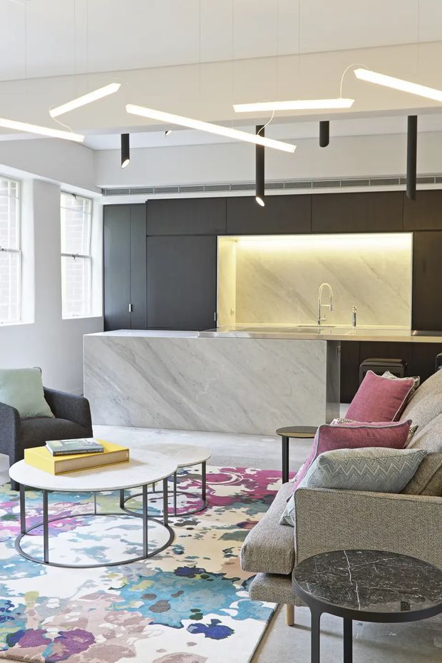 Bright open-plan living room and kitchen with a contemporary fit-out. The space features dark full-height kitchen cabinetry, a large pale marble-look island, illuminated stone splashback, white walls and ceiling, tall windows, sculptural linear pendant lighting, a textured neutral sofa with colourful cushions, round nesting coffee tables, and a large multicoloured abstract rug.