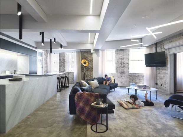 A bright open-plan loft-style living space with a polished concrete floor, exposed white-painted structural beams, white-painted brick walls, large industrial windows, and a minimalist kitchen with a large pale stone island. The room combines a lounge area, kitchen, and casual dining zone, with modern linear pendant and track lighting, dark upholstered seating, and a wall-mounted television.