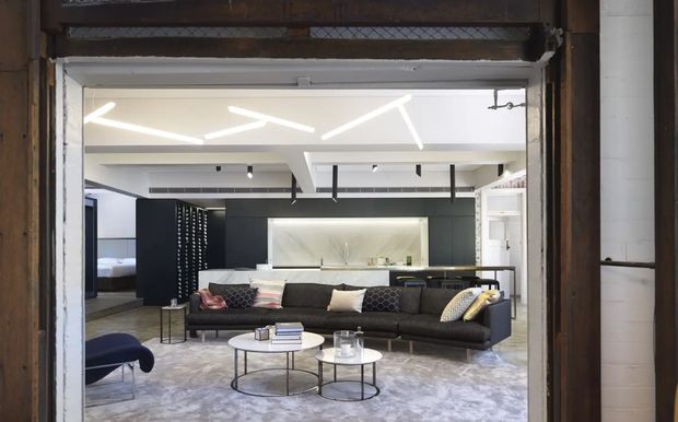 A contemporary open-plan living room viewed through a wide opening, with a large dark sectional sofa, round nested coffee tables, patterned area rug, sculptural linear ceiling lighting, and a sleek kitchen or bar area behind. Dark cabinetry and shelving contrast with white walls and ceiling, while the surrounding opening retains exposed dark timber framing and an industrial character.