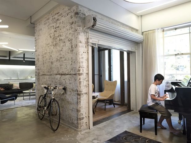 Open-plan loft-style interior with a raw exposed brick structural column, polished concrete flooring, and a partially enclosed music area containing a black grand piano. A child is playing the piano beside a bicycle, while modern furniture and a kitchen or living area are visible in the background. Large windows with curtains provide natural light, and the ceiling includes recessed and linear lighting.