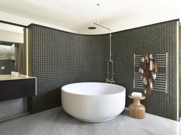 Contemporary bathroom featuring a freestanding white oval bathtub, wall-mounted shower hardware, dark textured grid-pattern wall tiles, pale stone-look flooring, a floating dark vanity, and a heated towel rail. The curved tiled feature wall creates a dramatic, cocoon-like setting, while slim black framing and warm vertical lighting add contrast.