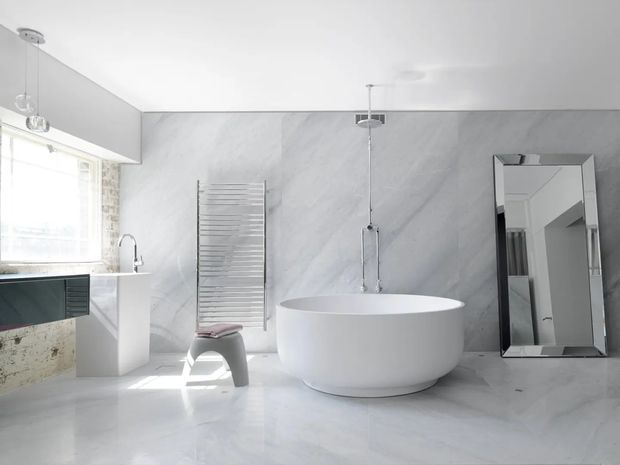 Bright contemporary bathroom with a freestanding white oval bathtub, wall-mounted rain shower, heated towel rail, floating vanity with basin, and large-format pale grey marble-effect wall and floor surfaces. A full-height mirrored panel leans against the wall, while daylight enters through the window on the left.