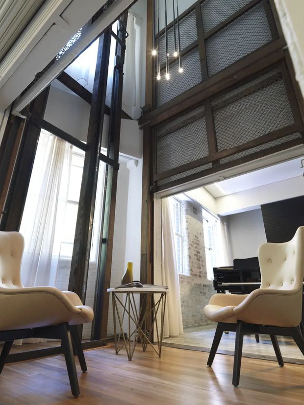 A tall, light-filled interior with polished timber flooring, two cream upholstered armchairs, and a small metal-framed side table. Dark timber-framed glazing and doors divide the room from a piano area, while an industrial-style mezzanine or upper-level walkway with dark framing and wire mesh overlooks the space. Exposed brick, high ceilings, and pendant lights add architectural character.