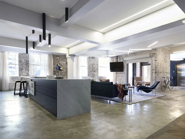 Open-plan loft-style interior with a polished concrete floor, exposed white-painted brick walls, structural beams, and large industrial windows. A dark kitchen island with a pale stone waterfall end anchors the foreground, while a lounge area with dark sofas, a wall-mounted television, sculptural seating, bicycles, and black linear pendant lights occupies the background. The space is bright, spacious, and industrial-contemporary.