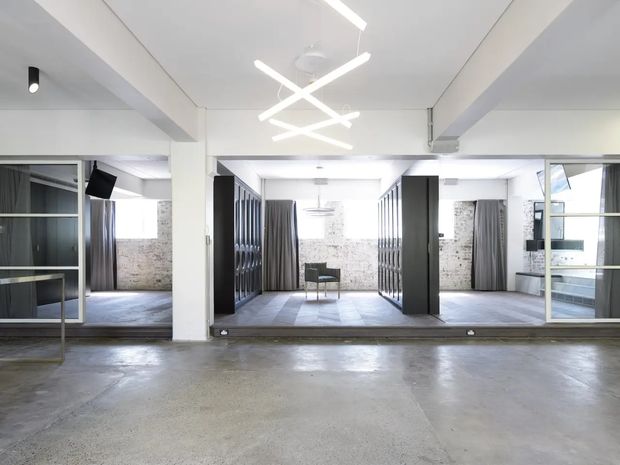 A bright, open-plan commercial or studio interior with polished concrete flooring, exposed white-painted structural beams, extensive black-framed glass partitions, dark vertical storage or display joinery, and exposed light brick walls. Grey curtains divide several work or meeting areas, while linear white pendant lights create a contemporary industrial atmosphere.