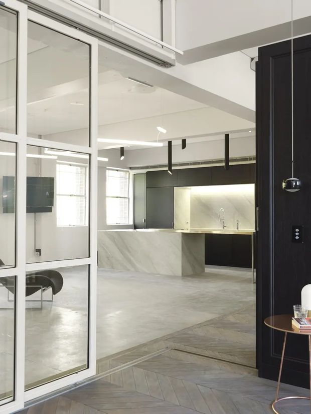 A bright, open-plan contemporary kitchen and living area viewed through a glazed steel-framed partition and an open dark timber door. The space features a large marble-look island and backsplash, dark fixed cabinetry, polished concrete flooring in the main room, and a contrasting geometric timber parquet floor in the foreground. Linear and pendant lighting, exposed structural beams, and expansive internal glazing contribute to an industrial-modern fit-out.