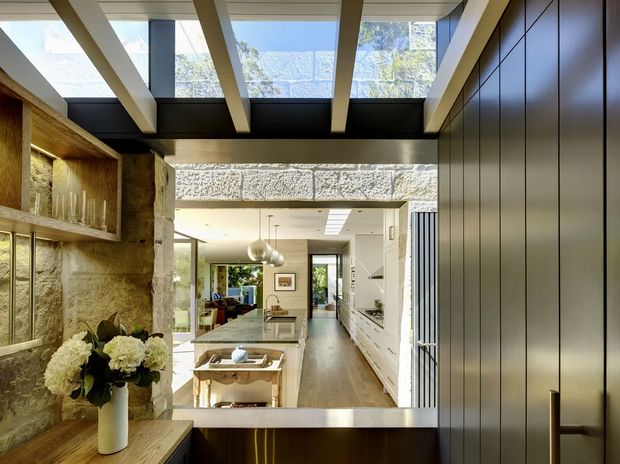 A refined contemporary interior viewed from a darker entry or circulation zone toward an open-plan kitchen and living area. The space combines exposed rough stone walls, a long white kitchen with integrated cabinetry, warm timber flooring, dark full-height joinery, and a dramatic overhead structure of deep beams with clerestory glazing. A sculptural pendant hangs above the kitchen island, while a framed view extends through the kitchen toward the living room and greenery beyond.