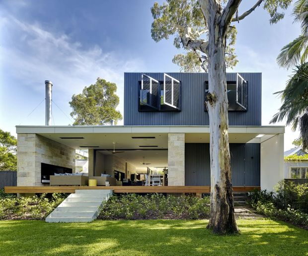 Front exterior view of a contemporary two-storey residence with a dark vertically ribbed upper volume, broad flat-roofed covered terrace, pale masonry columns, timber decking, louvred window shutters, and landscaped lawn. The open ground floor reveals an outdoor living area and glimpses of the interior.