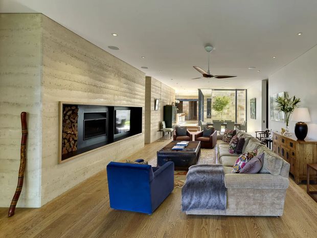 A spacious contemporary living room with a long textured pale stone or concrete feature wall incorporating a built-in fireplace, television and firewood storage. The room has warm timber flooring, a large light-grey sectional sofa, blue and brown lounge chairs, a dark coffee table, and timber cabinetry. Floor-to-ceiling glazing at the rear brings in natural light and connects the room to an outdoor area. A ceiling fan and recessed lighting complete the relaxed, refined interior.