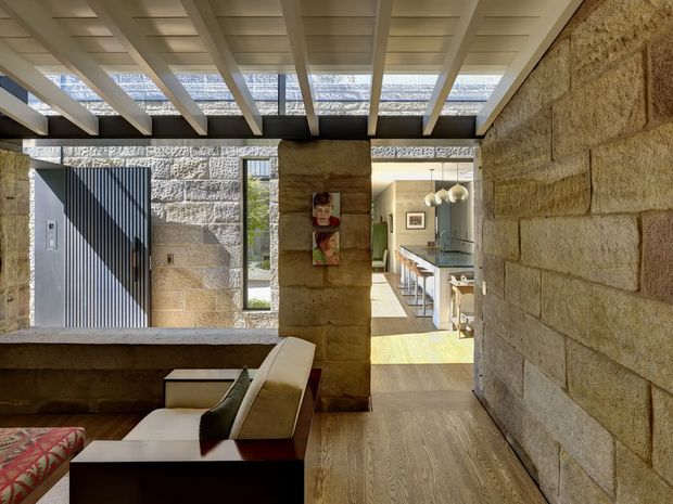 Interior view of a contemporary open-plan home with exposed rough-cut stone masonry walls, a timber-look floor, and a striking exposed roof structure with white beams and translucent overhead panels. The foreground contains a partially visible living area with built-in seating and a low dark table, while a dining and kitchen area is visible beyond the central stone pier. Strong natural light filters through the roof, creating patterned shadows and a warm, textural atmosphere.