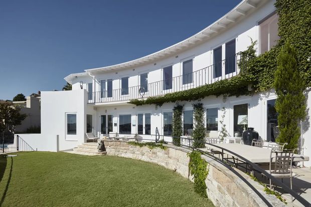 Exterior view of a large white two-storey residence with a gently curved facade, extensive glazing, upper-level balcony with slim metal balustrade, and climbing greenery along the terrace edges. A curved stone retaining wall borders a manicured lawn and paved outdoor terrace with dining furniture. The bright, refined design has coastal and Mediterranean influences.