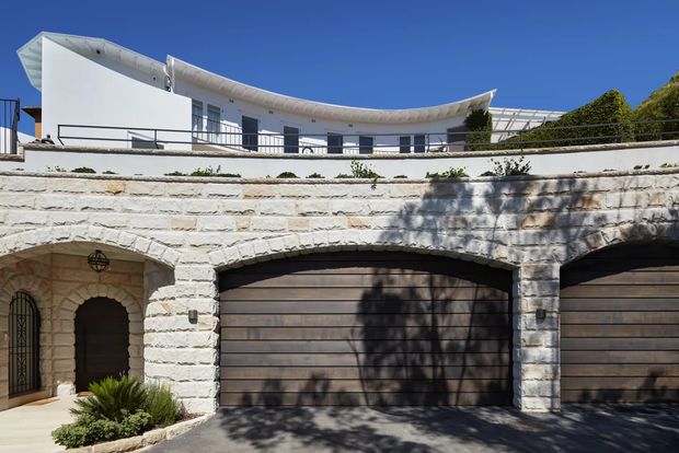Exterior view of a multi-level residence with a curved white upper-storey facade and sweeping roofline, dark metal balcony railings, and a substantial lower level clad in pale rough-cut stone. The lower facade contains multiple wide timber-toned garage doors and an arched pedestrian entry, with landscaped planting at the driveway edge. Bright clear daylight emphasizes the contrast between the white rendered upper level, stonework, and dark garage doors.