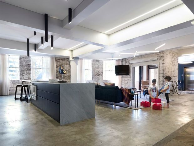 Bright open-plan loft-style interior combining a kitchen, living area and dining space. The room features polished concrete flooring, exposed white-painted brick walls, large industrial windows, a dark island with a pale stone waterfall side, black linear pendant lights, exposed structural beams and a high ceiling. A dark sofa, television and casual furnishings occupy the living area, with bicycles and children's toys visible near the rear entry.