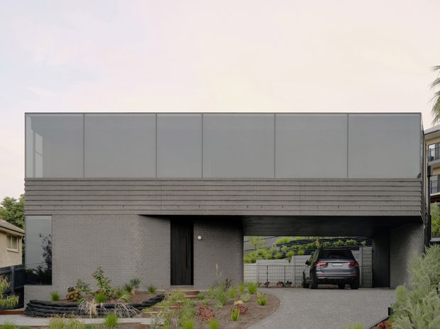 Street-facing view of a contemporary two-storey residence with a minimalist grey masonry lower level, broad horizontal upper cladding panels, a recessed dark entry door, and an integrated covered carport. A planted front garden and gravel driveway occupy the foreground, with neighbouring homes visible at the sides.