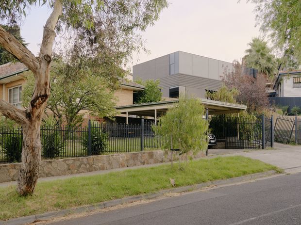 Street-facing view of a contemporary multi-level residential property in a leafy suburban setting. The home combines a boxy upper volume clad in grey horizontal and smooth panels with lower pitched-roof brick sections, screened by mature trees and landscaping. A broad covered carport, black metal fencing, front garden, driveway, and stone retaining wall are visible.