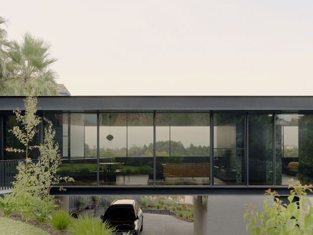 A contemporary two-storey residential building with a long, low roofline and extensive floor-to-ceiling dark-framed glazing. The upper level appears to contain an open-plan living or dining space, visible through the glass, while the lower level forms a sheltered carport and landscaped entry. The design features strong horizontal lines, dark metal framing, reflective glazing, and integrated garden planting.
