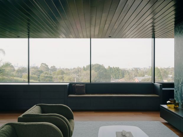 A calm, contemporary living room with expansive floor-to-ceiling windows overlooking a leafy suburban landscape. The room features a long dark built-in window seat or sofa, curved upholstered lounge chairs, dark wall panelling and cabinetry, a warm timber floor, a pale rug, and a linear slatted ceiling. The restrained palette and panoramic glazing create an airy yet moody interior.