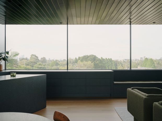 A refined contemporary living space with expansive floor-to-ceiling glazing overlooking leafy suburban surroundings. Dark built-in cabinetry runs beneath the windows, paired with a dark stone island or counter in the foreground, a low-profile dark sofa, and warm timber flooring. The ceiling is clad in narrow linear timber panels, creating a strong horizontal rhythm and a calm, sophisticated atmosphere.