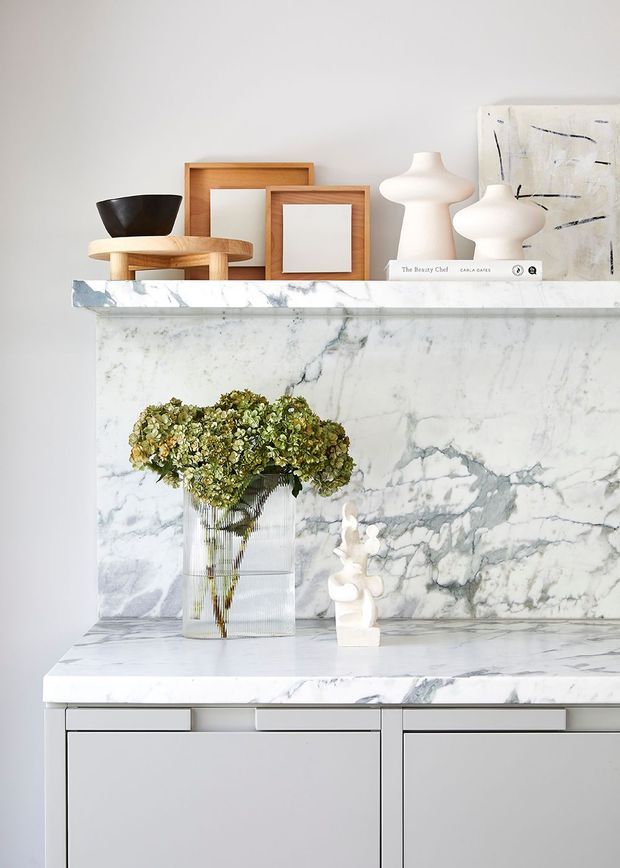 A refined interior vignette featuring a white and grey-veined marble fireplace or built-in display surround with a matching projecting shelf and base. Minimal pale cabinetry is visible below, while the shelf is styled with timber-framed artwork, sculptural white ceramics, a dark bowl, books, and a vase of dried greenery. The look is bright, calm, and contemporary with strong natural-stone veining.