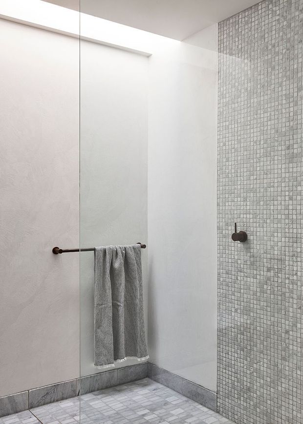 Minimalist walk-in shower with a frameless glass screen, pale textured plaster walls, grey mosaic-tiled feature wall and floor, and dark bronze shower and towel hardware. The space is bright, restrained and contemporary.