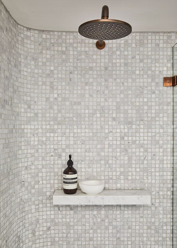 A compact walk-in shower with small square pale stone mosaic tiles covering the curved walls. A dark bronze ceiling-mounted rainfall shower and matching wall fitting contrast with the light tiled enclosure. A simple white stone shelf holds a soap dispenser and bowl, creating a clean, refined bathroom detail.