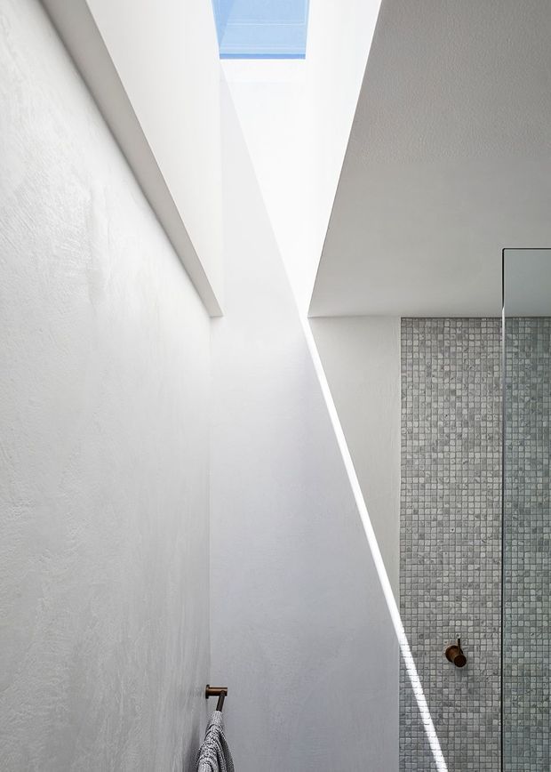 A bright, minimalist bathroom interior featuring a walk-in glass shower enclosure, small grey mosaic tile wall, white textured plaster walls, and a dramatic skylight that casts a strong diagonal shaft of light. A dark bronze shower fitting and towel rail provide subtle contrast.