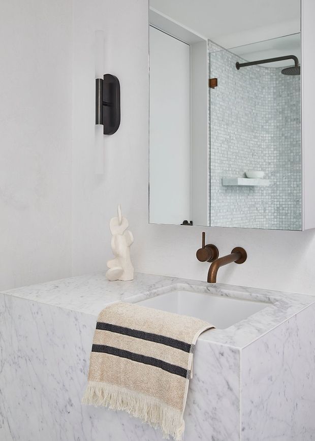 A refined bathroom vanity with a white marble-clad basin, wall-mounted bronze tapware, a large mirror, and a walk-in shower visible in the reflection. The shower features small pale mosaic tiles, a dark bronze rainfall showerhead and arm, and a floating shelf. The space has white walls, minimalist lighting, and a neutral palette with subtle black and bronze accents.