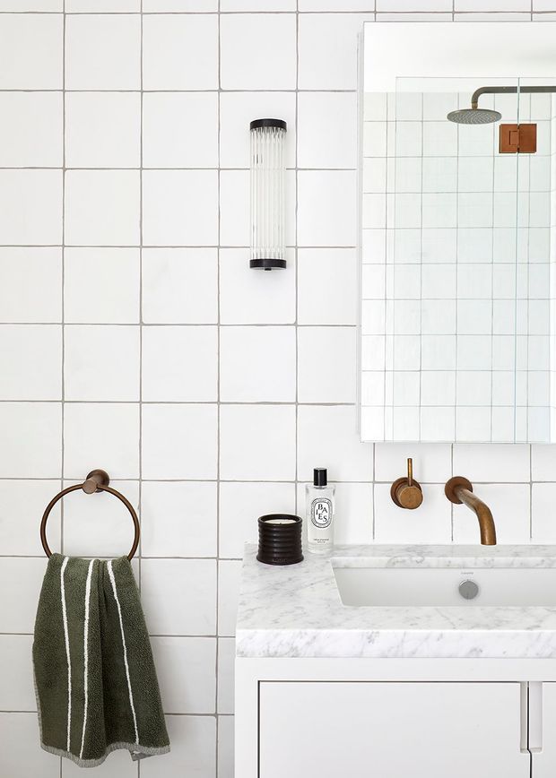 A compact, bright bathroom vanity area with white square wall tiles, a white marble-look benchtop, integrated white basin, and white built-in cabinetry. A round bronze towel ring with a dark green towel, bronze wall-mounted tapware, a large mirror, and a cylindrical wall light add warm contrasting accents. The shower is visible in the mirror reflection.