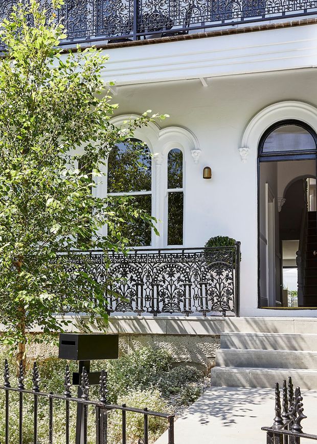 Front exterior of a refined heritage-style residence, featuring a white rendered facade, arched windows and doorway, ornate black wrought-iron balcony and boundary railings, broad stone steps, and a landscaped garden entry. The open doorway reveals a dark interior hall and staircase.