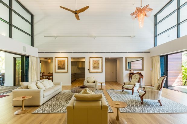 A bright, expansive living room with a double-height ceiling and extensive floor-to-ceiling glazing. The space features pale timber flooring, white walls and ceiling panelling, large cream sofas, patterned area rugs, sculptural timber side tables, classic upholstered armchairs, built-in shelving, framed artwork, and distinctive suspended pendant lights and ceiling fans. The open-plan room has a refined contemporary character with warm mid-century-inspired furnishings.