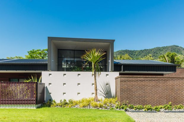 Front exterior view of a contemporary single-storey residence with a low-pitched dark metal roof, projecting framed entry or upper glazing volume, white rendered wall with small square openings, and extensive brown brickwork. A timber slat gate, landscaped garden, and mountainous vegetation form the foreground and background.