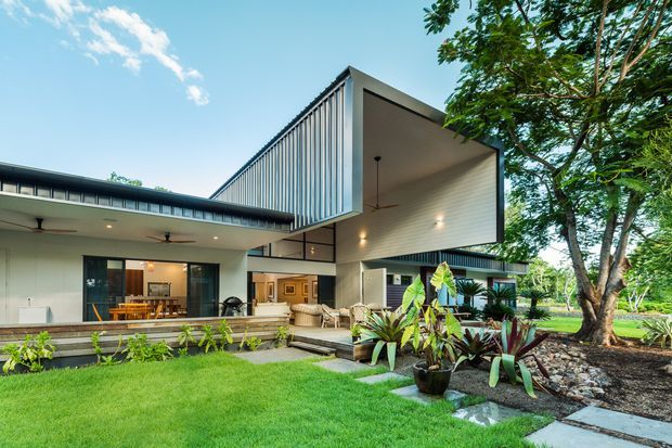 Exterior view of a contemporary two-storey residence with strong geometric forms, a projecting upper-level volume, vertical facade screening, broad covered outdoor areas, and extensive glazing. The home opens onto a landscaped lawn and planted garden with a paved walkway and mature trees.