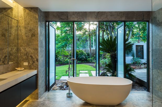 Contemporary bathroom with a freestanding white oval bathtub positioned in front of full-height black-framed glazed doors opening to a lush private garden. The room features dark grey stone-look wall and floor finishes, a floating dark vanity with a pale benchtop, a wall-mounted mirror, and minimalist fittings. The open glazing brings abundant greenery and natural light into the space.