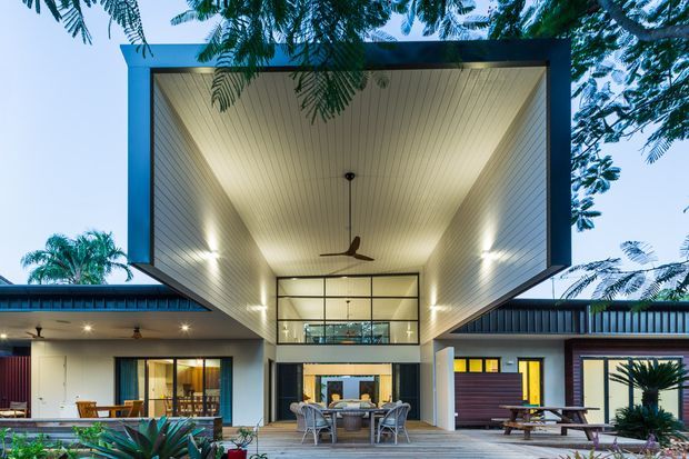 Exterior view of a contemporary single-storey residence centred on a dramatic projecting, box-like roof canopy over an open outdoor dining and living area. The house features extensive floor-to-ceiling glazing, dark-framed windows and doors, pale horizontal wall cladding, warm timber-lined soffits, integrated recessed lighting, and a central double-height glazed volume. A timber deck, outdoor dining furniture, tropical planting, and surrounding mature trees create a sheltered indoor-outdoor living setting.