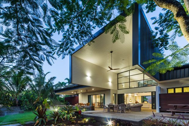 Exterior architectural photograph of a contemporary, multi-level home set within lush tropical landscaping. The house features a dramatic projecting roof canopy with a light timber-lined soffit, dark vertical cladding, extensive floor-to-ceiling glazing, a covered outdoor living and dining area, broad timber decking, and integrated exterior lighting. A double-height glazed volume and sculptural roof form create a strong indoor-outdoor connection.