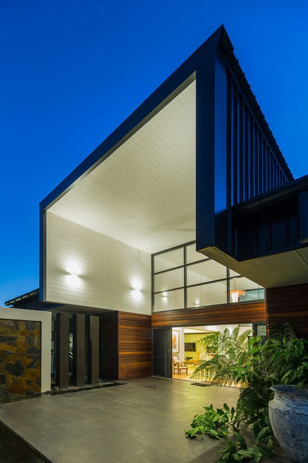 Dusk exterior view of a modern multi-level residence with a dramatic angular roof and deep white-lined cantilevered soffit. Dark vertical metal screening and exposed structural framing contrast with warm horizontal timber cladding and extensive glazed openings. A paved entry courtyard, stone feature wall, planter, and lush tropical planting lead toward a brightly lit interior.