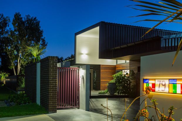 Twilight exterior photograph of a contemporary single-storey residence. The house features strong geometric volumes, a projecting gable-like upper form clad in dark vertical metal, timber-look wall cladding, exposed concrete surfaces, a dark brick boundary wall, a slatted entry gate, and colourful illuminated glazing or artwork beside the entry. Landscape planting and a palm frame the modern facade.