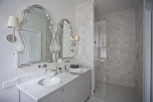 Elegant traditional bathroom featuring a double vanity with a white marble-look benchtop, twin sculptural framed mirrors, wall sconces, pale grey cabinetry, and a tiled walk-in shower enclosed by clear glass. The space uses soft neutral finishes and decorative detailing for a refined, classic look.