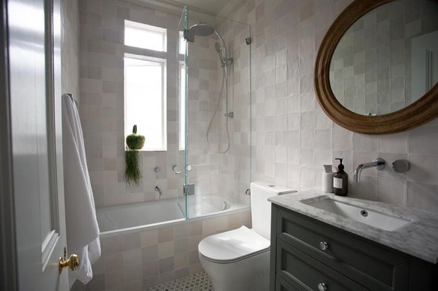 A bright, compact traditional bathroom with a white tiled tub and shower enclosure, freestanding toilet, and dark painted vanity topped with pale marble or stone. A large round wood-framed mirror, brass door hardware, chrome fixtures, and a small window add classic character.